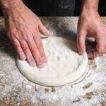 in this image men is pressing the pizza dough on a flat surface